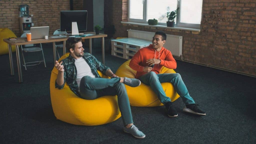 Two men sit in yellow beanbag chairs in an industrial-style office, talking and holding game controllers.