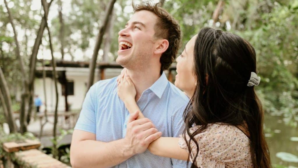 A woman in a floral dress places her hand on the neck and arm of a laughing man.