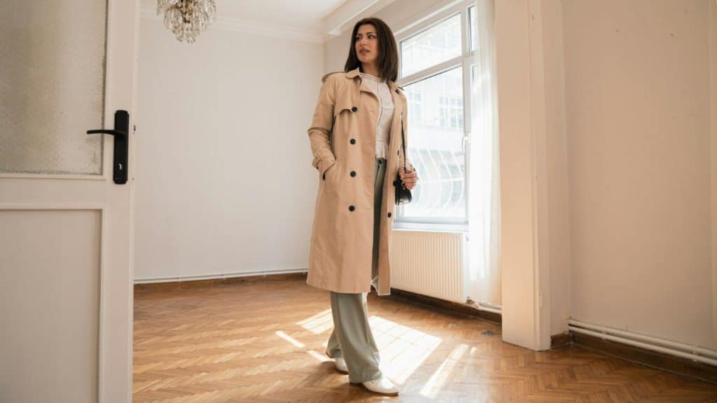 A woman in a tan trench coat stands in an empty room looking toward a window.