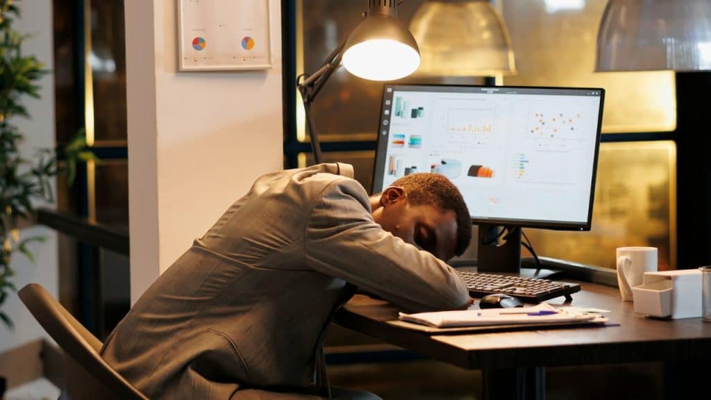 A man in a grey suit rests his head on a desk in front of a computer.