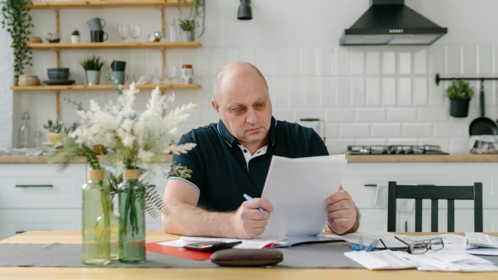 A man sits at a kitchen table reviewing papers with a pen and calculator nearby.