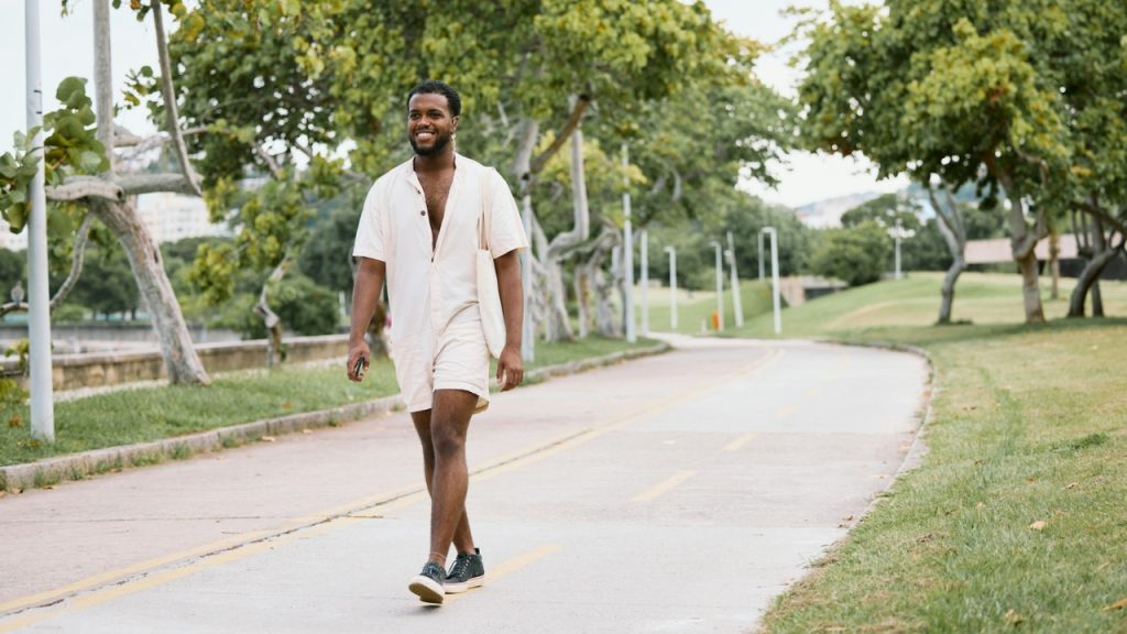 A smiling man in a light-colored outfit walks along a paved path lined with trees.