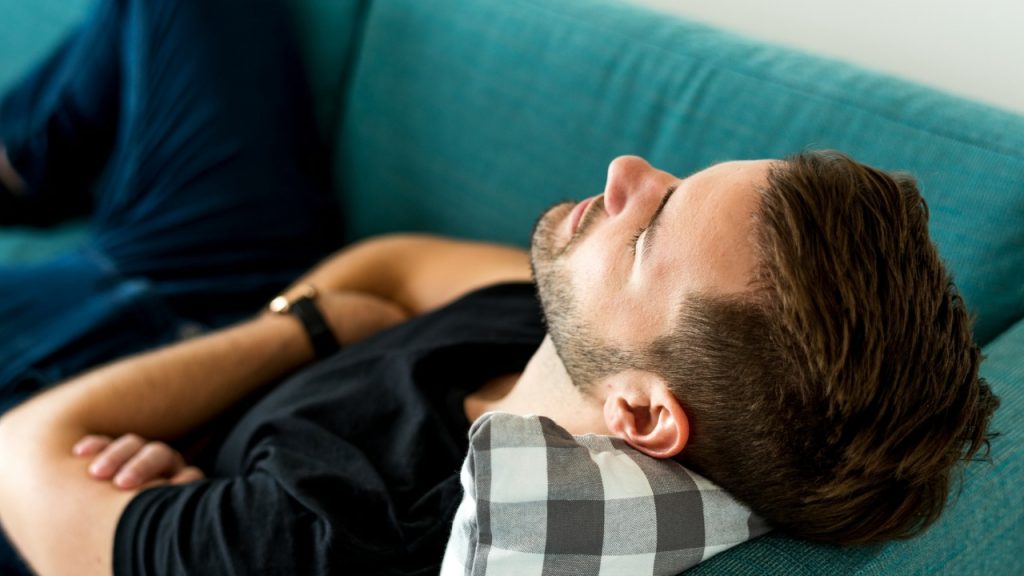 A man with his eyes closed lies on a teal sofa with his arms crossed.