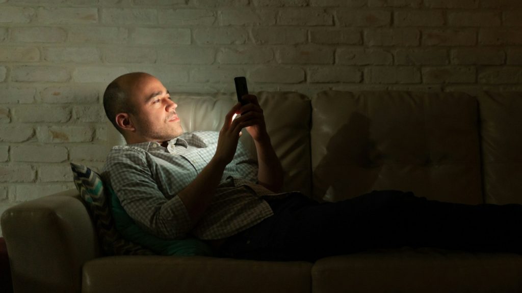 A man lies on a sofa in a dark room looking at his phone.
