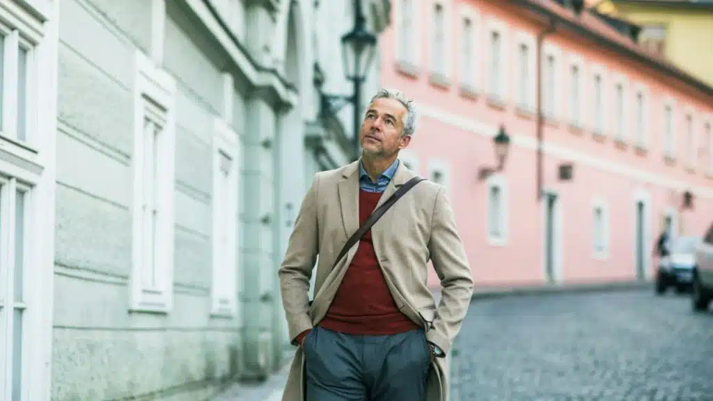 A man in a tan coat and red sweater walks along a cobblestone city street.