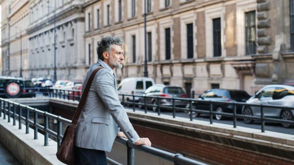 A bearded man in a suit jacket stands on a bridge, looking out at a city street.