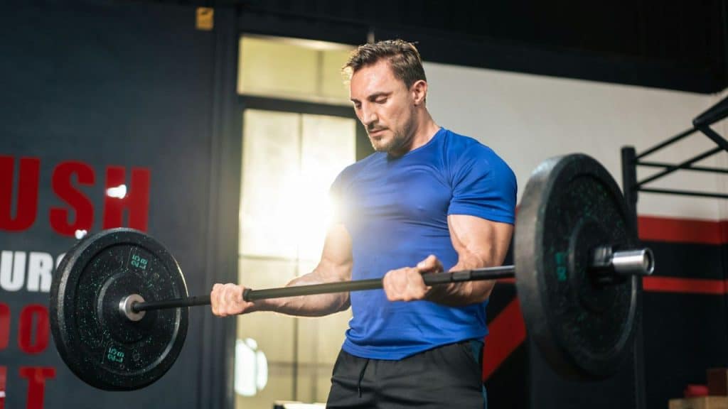 A muscular man in a blue t-shirt performs a bicep curl with a weighted barbell.