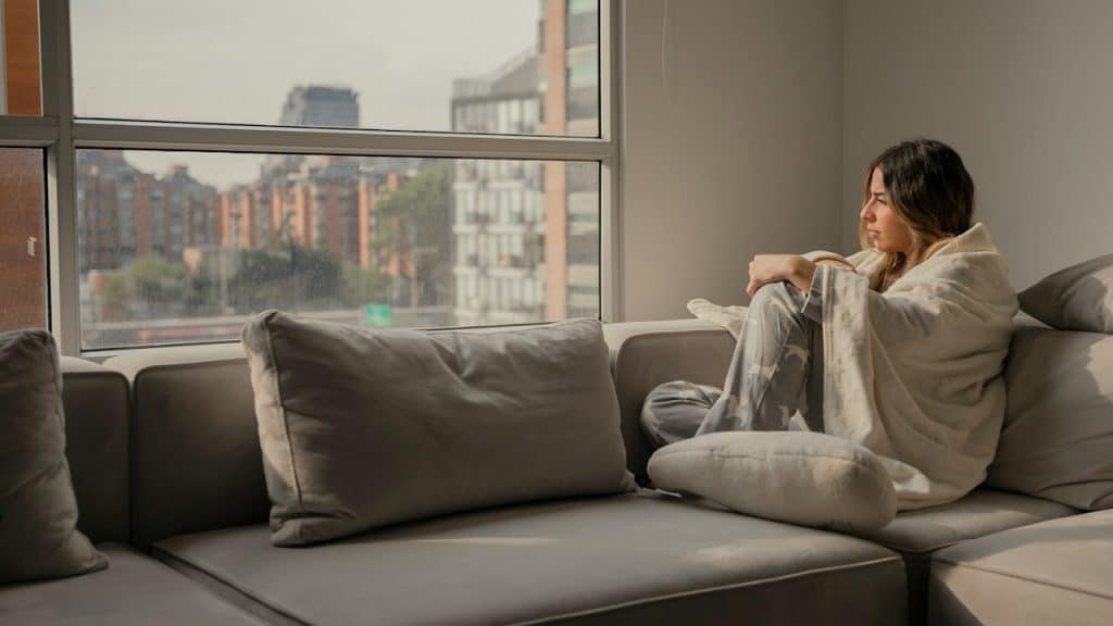 A woman sitting on a couch covered in a sheet and looking out of a window at the urban skyline.