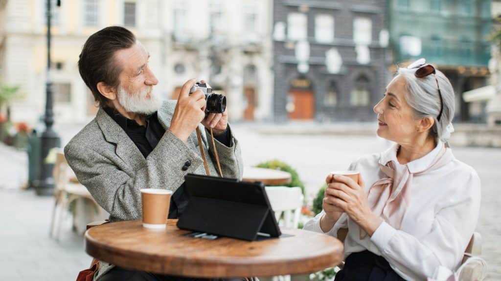 An older man takes a photo of a woman at an outdoor cafe table.