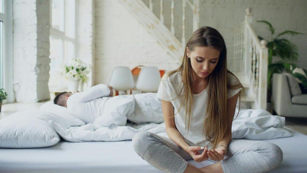 A woman sitting on a bed looking down while a man sleeps behind her.