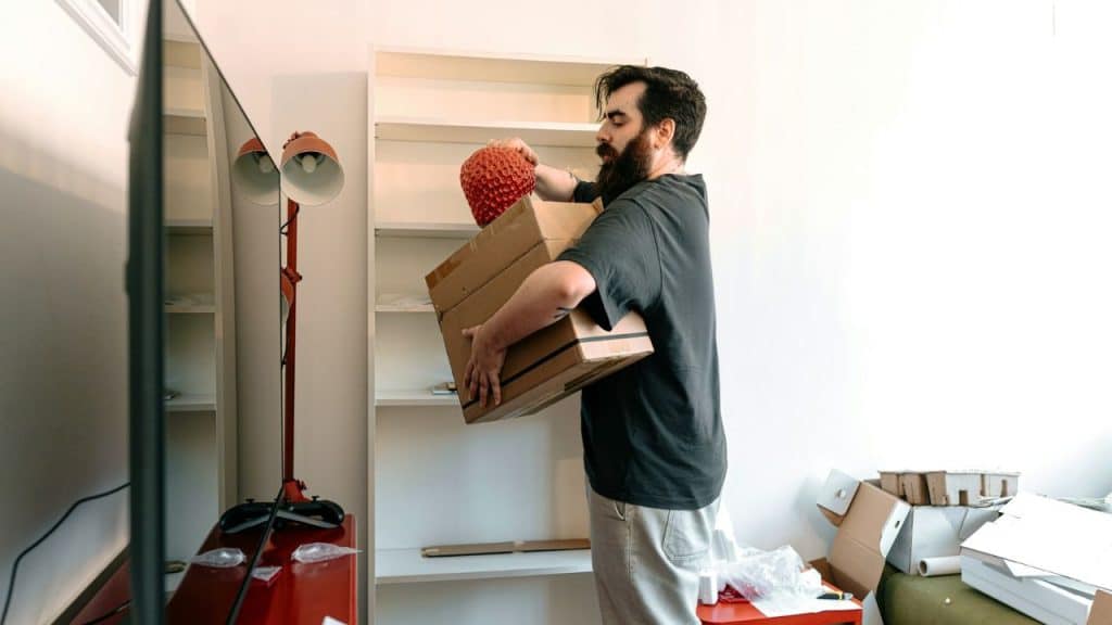 A bearded man carries a cardboard box and a red object near a white shelf.