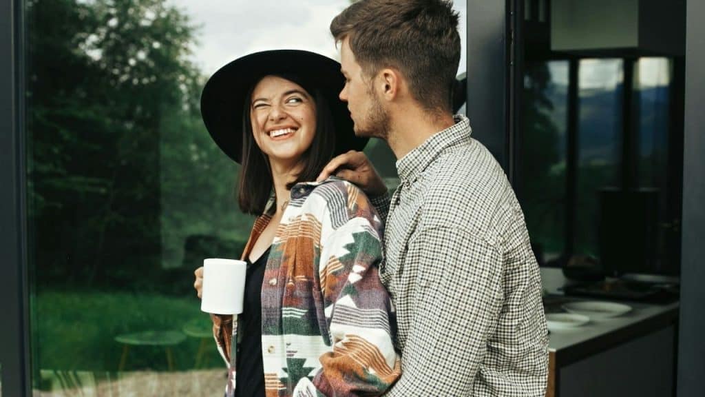 A laughing woman in a black hat holds a mug while a man leans in.