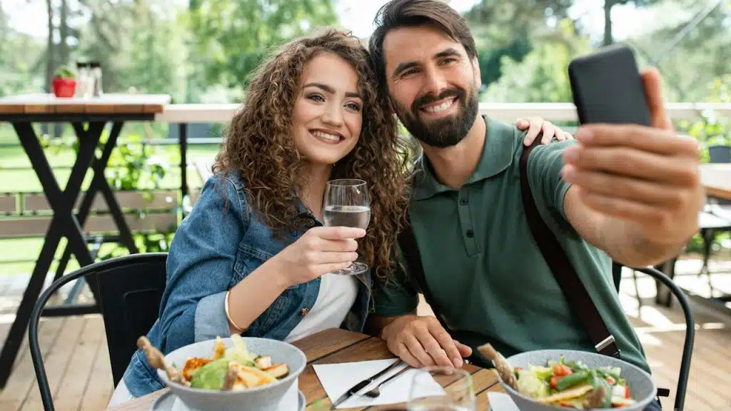 Happy couple sitting outdoors on terrace restaurant taking selfie with smartphone.