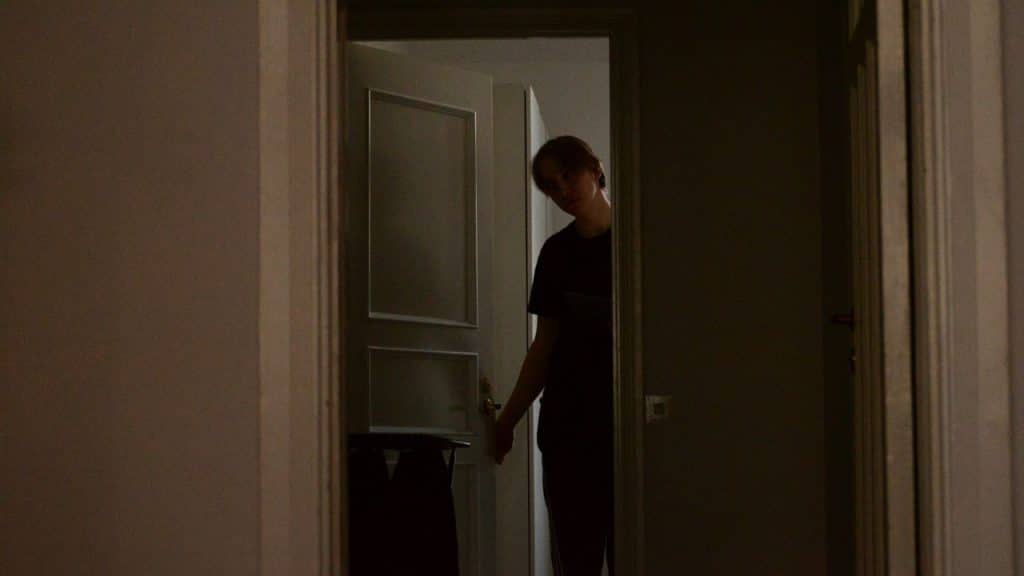 A person is seen from the shadows, opening a door and peering out.