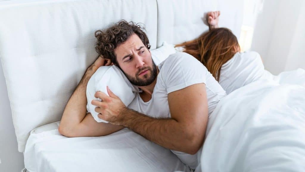 A man with curly hair lies in bed looking troubled while a woman sleeps behind him.
