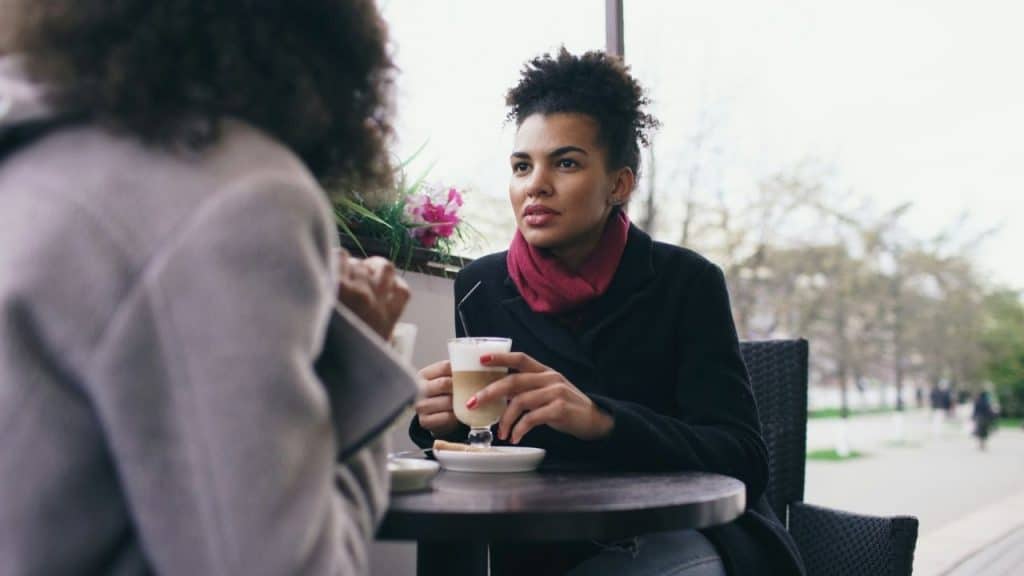 A woman with a red scarf holds a latte while looking at someone at a table.