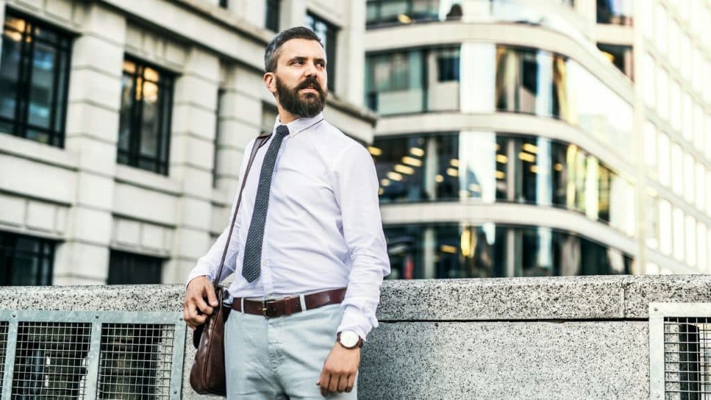 A bearded man in a white dress shirt and tie stands outdoors against a city building.