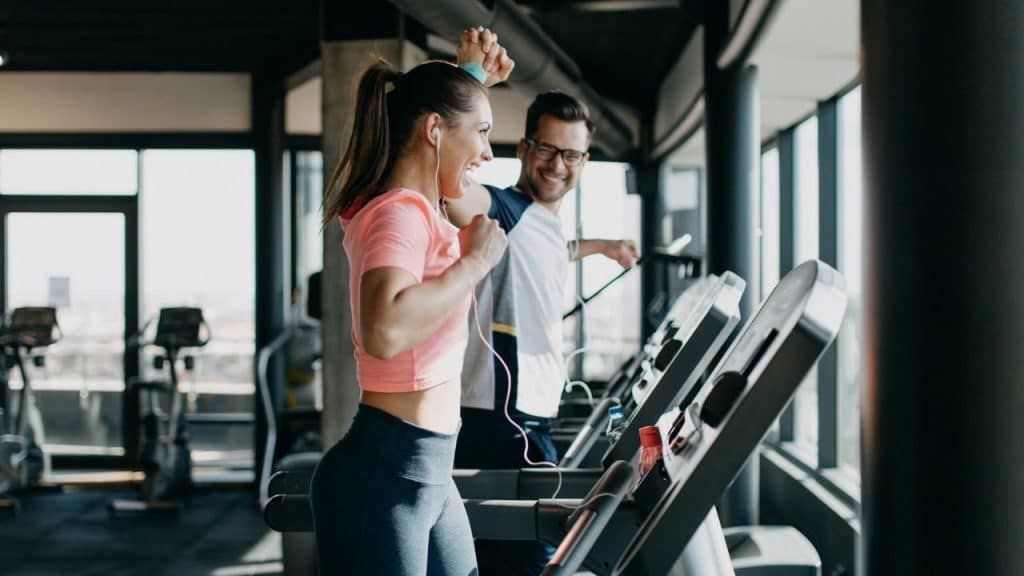 A smiling man and woman exercise on treadmills next to each other in a gym.