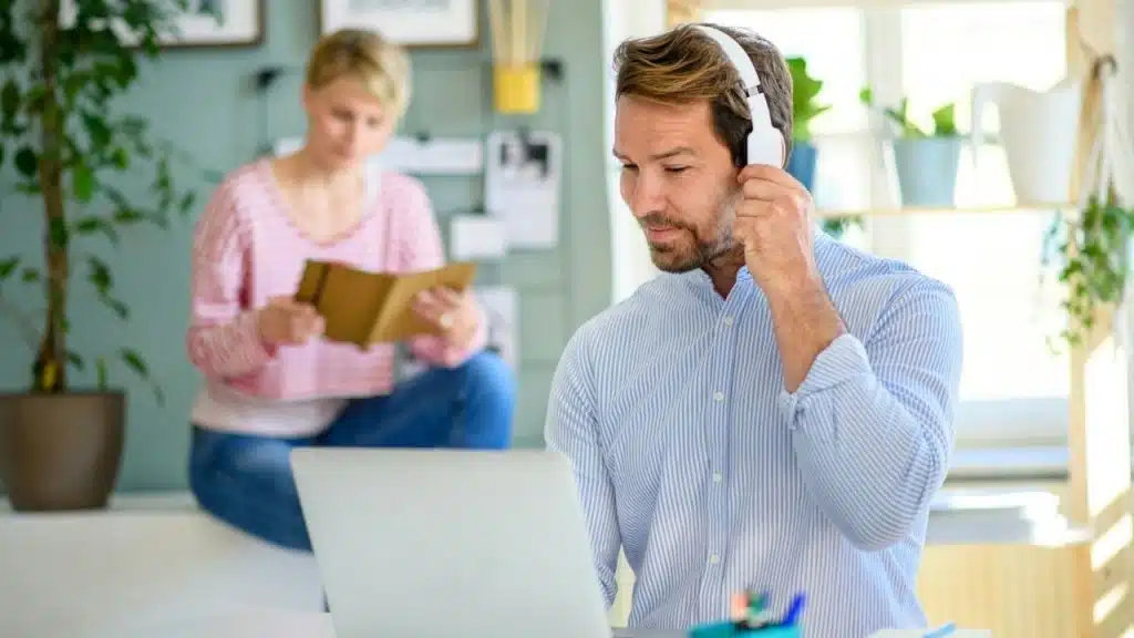 A man in a striped shirt wears headphones at a laptop while a woman reads behind him.