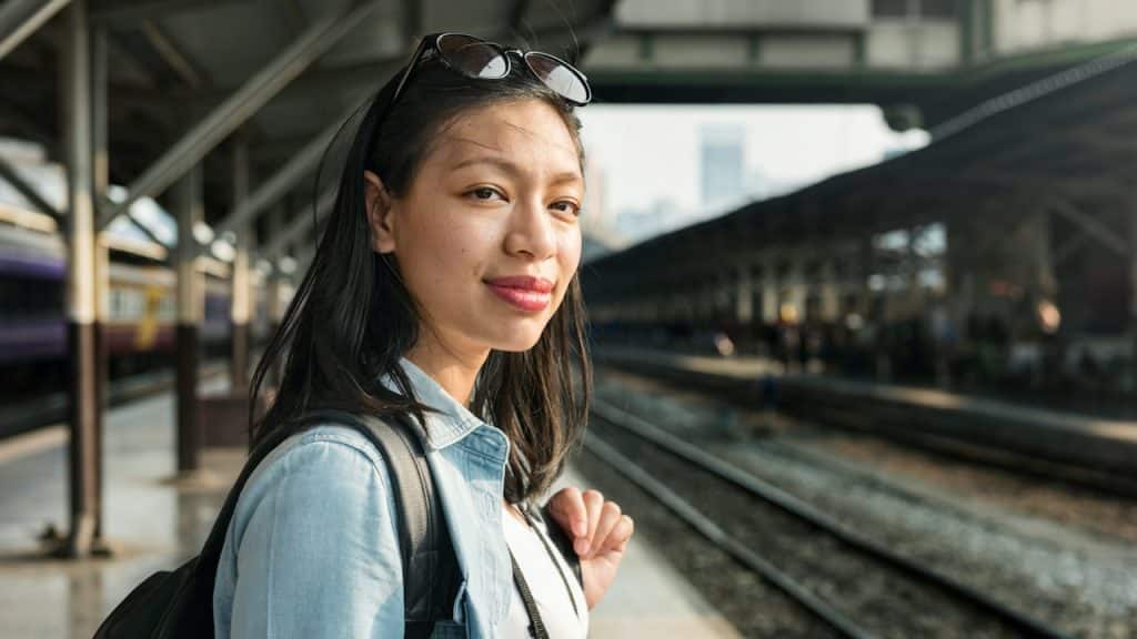 A young woman wearing casual jeans with a backpack is standing at a railway station.
