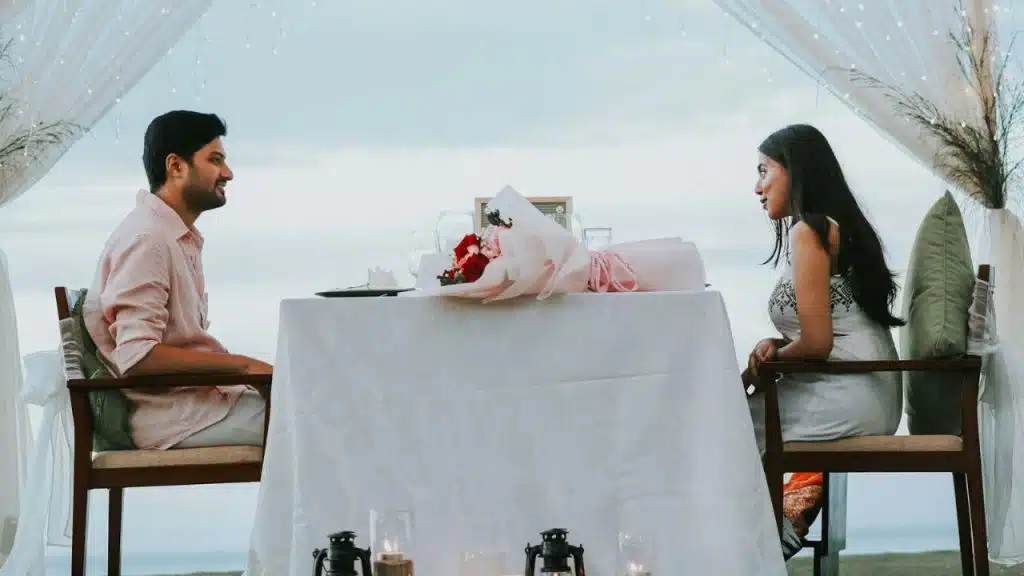 A man and woman sit facing each other at a table set for a meal outdoors.