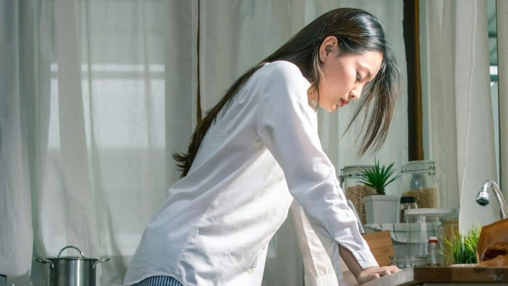 A woman in a white shirt stands in a kitchen leaning forward with her eyes closed.