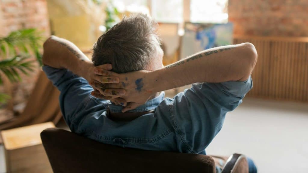 A man with paint-stained hands and tattooed arms rests his head in his hands indoors.