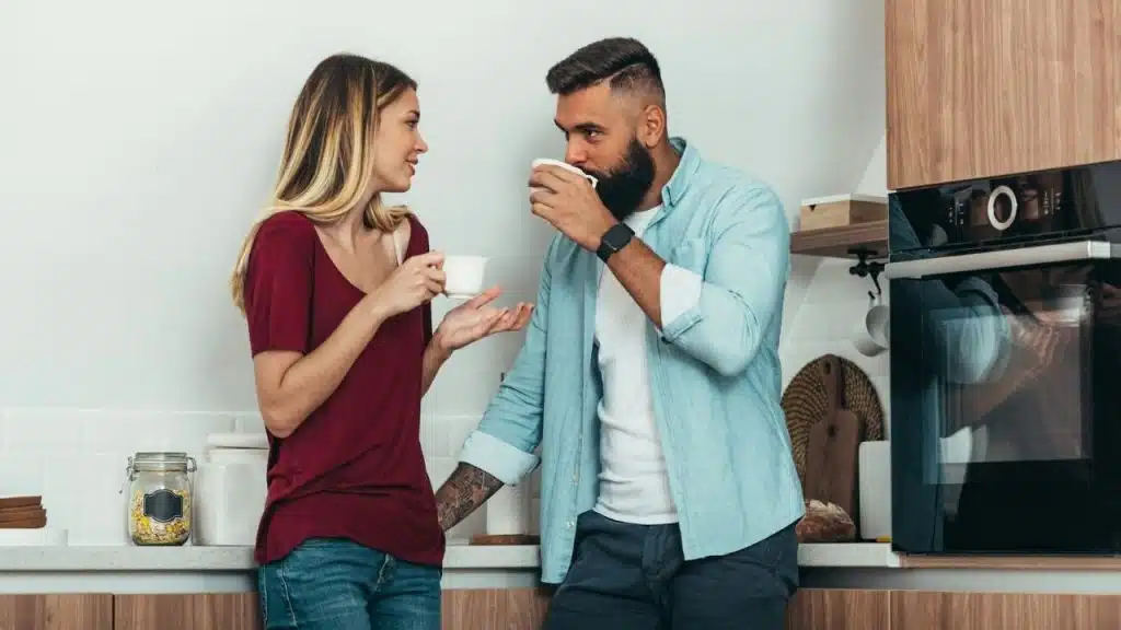 A man with a beard and a woman in a maroon shirt talk while drinking coffee.