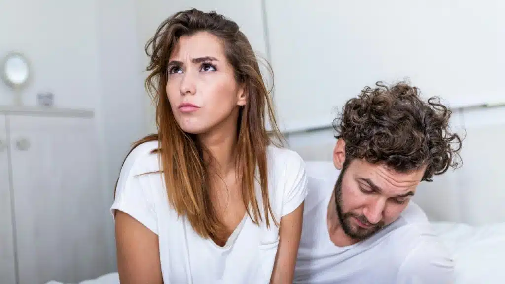 A woman looks upward with a frown while a man looks down beside her in bed.