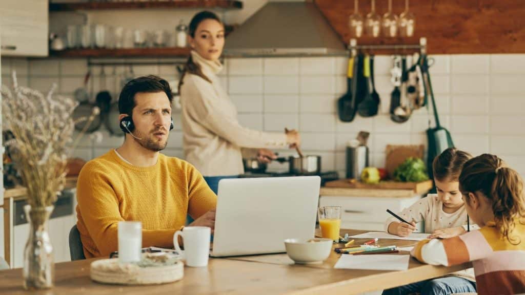 A man works at a laptop while a woman cooks and two children draw nearby.