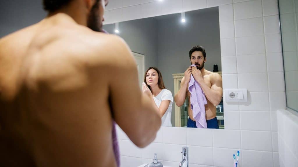 A shirtless man with a purple towel looks into a bathroom mirror beside a woman.