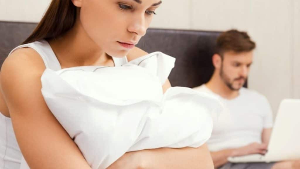 A woman hugs a pillow in the foreground while a man uses a laptop behind her.