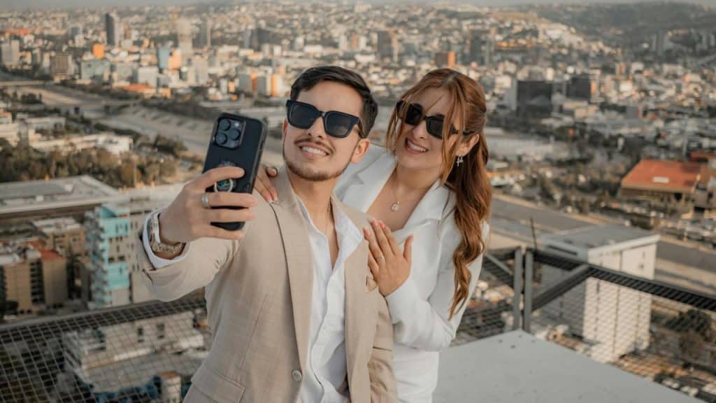 A stylish couple taking a selfie on a rooftop with a city skyline behind them.