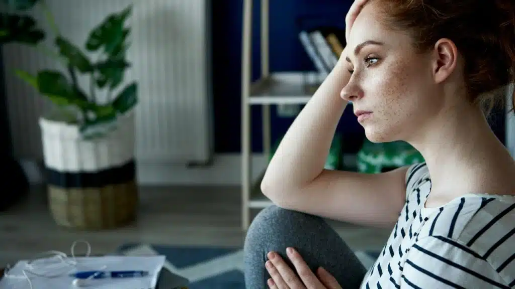 A person sitting indoors with one hand on their head, looking thoughtfully to the side.