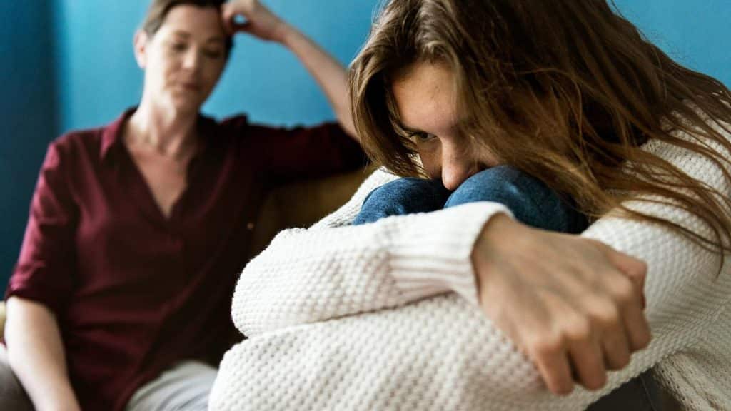 A distressed woman hugging her knees as another woman looks on.