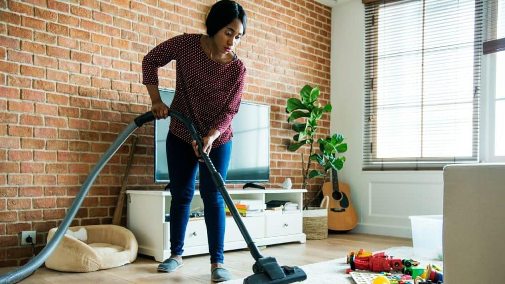 A woman vacuums a living room rug near scattered toys and a brick wall.