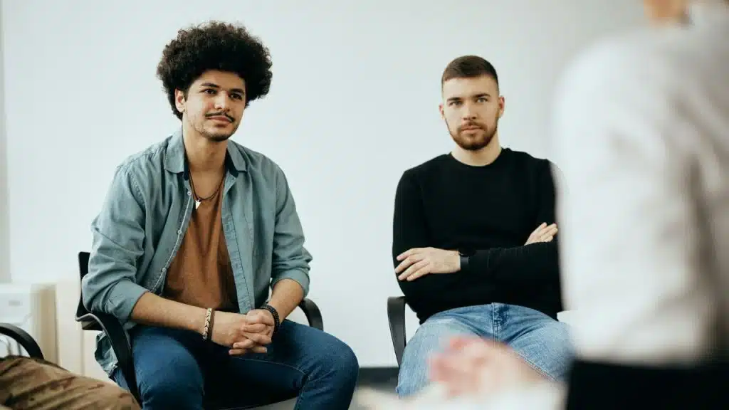 Two men sit in chairs during a meeting, looking toward a person standing in the foreground.