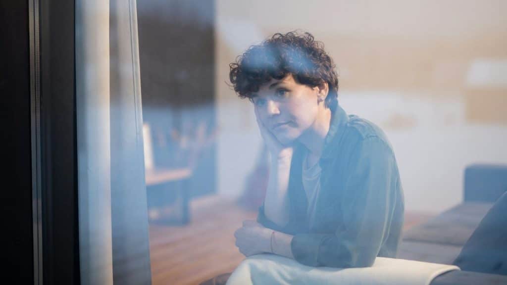 A woman with short curly hair rests her chin on her hand, looking out a window.