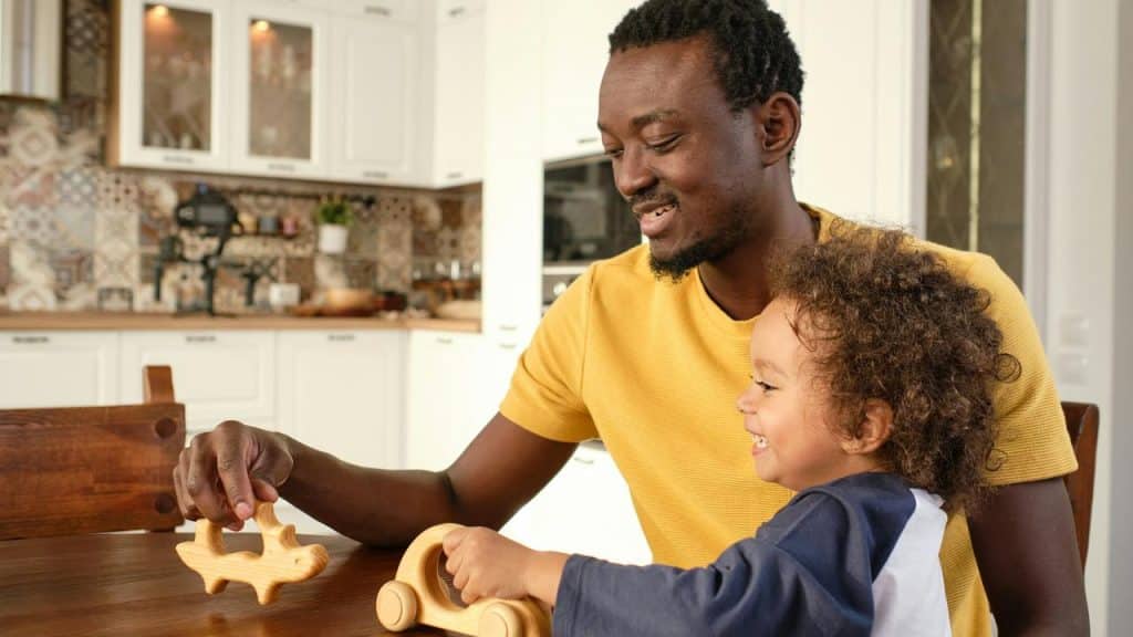 A man smiling as he plays with wooden toys alongside a young child at a kitchen table.