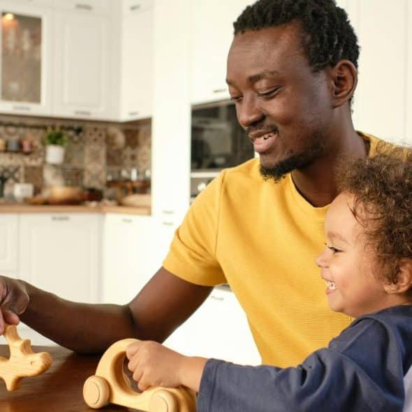 A man smiling as he plays with wooden toys alongside a young child at a kitchen table.