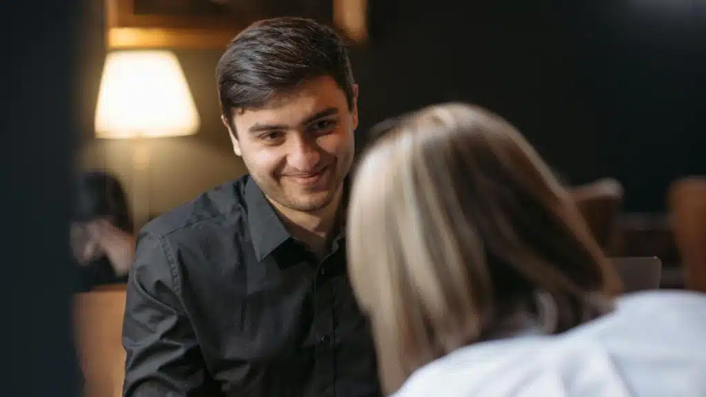 A man in a black shirt smirks while looking at a woman with blonde hair.