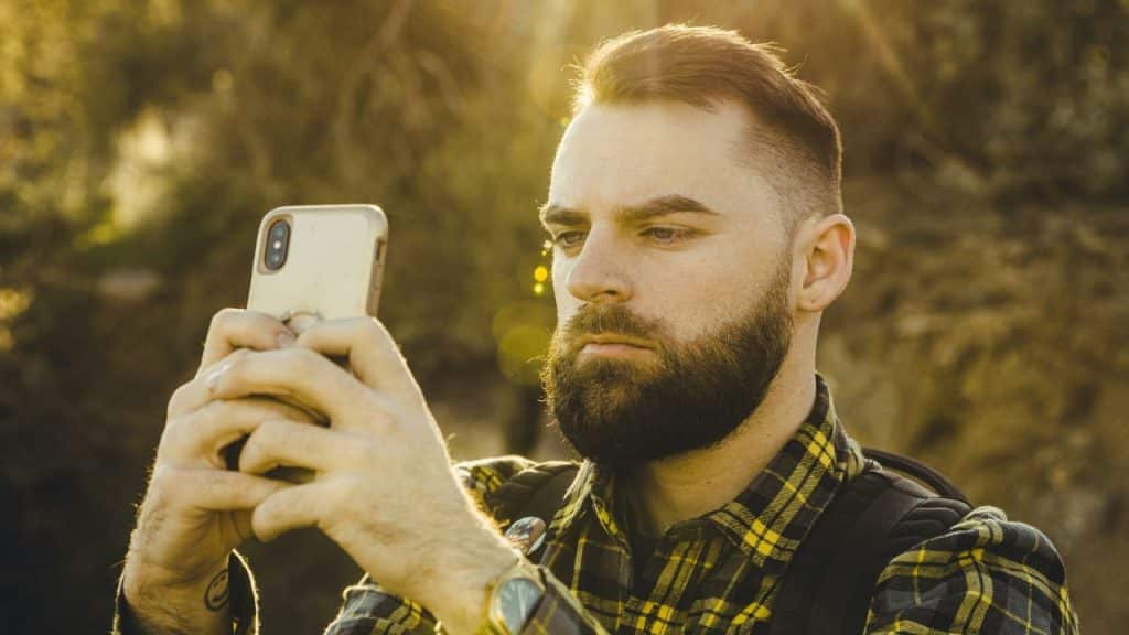 A bearded man in a plaid shirt holds up a smartphone outdoors during golden hour.