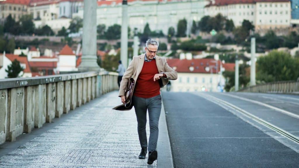 A man in a blazer walks across a bridge while looking down at his wristwatch.