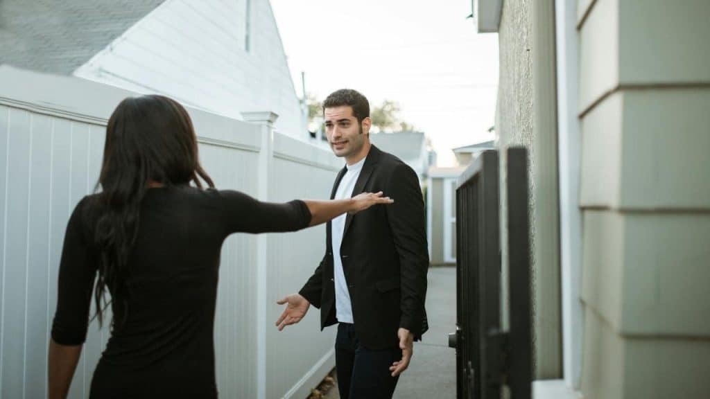 A man in a blazer talks with open hands to a woman in an outdoor walkway.