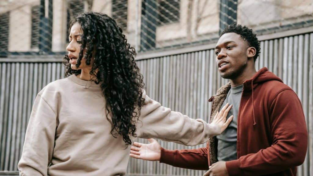 A woman with curly hair looks away as a man reaches out toward her chest.