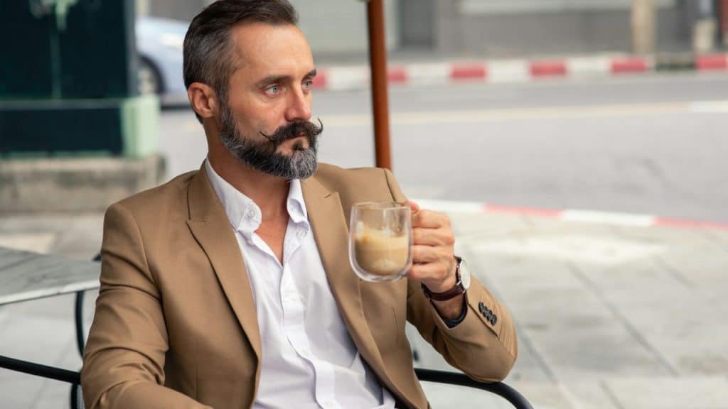 A man with a groomed mustache sits at an outdoor table holding a glass of coffee.