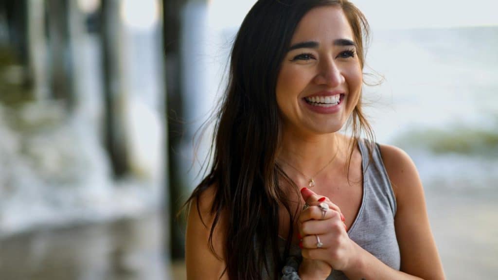 A smiling woman clasping her hands near a beachside pier.