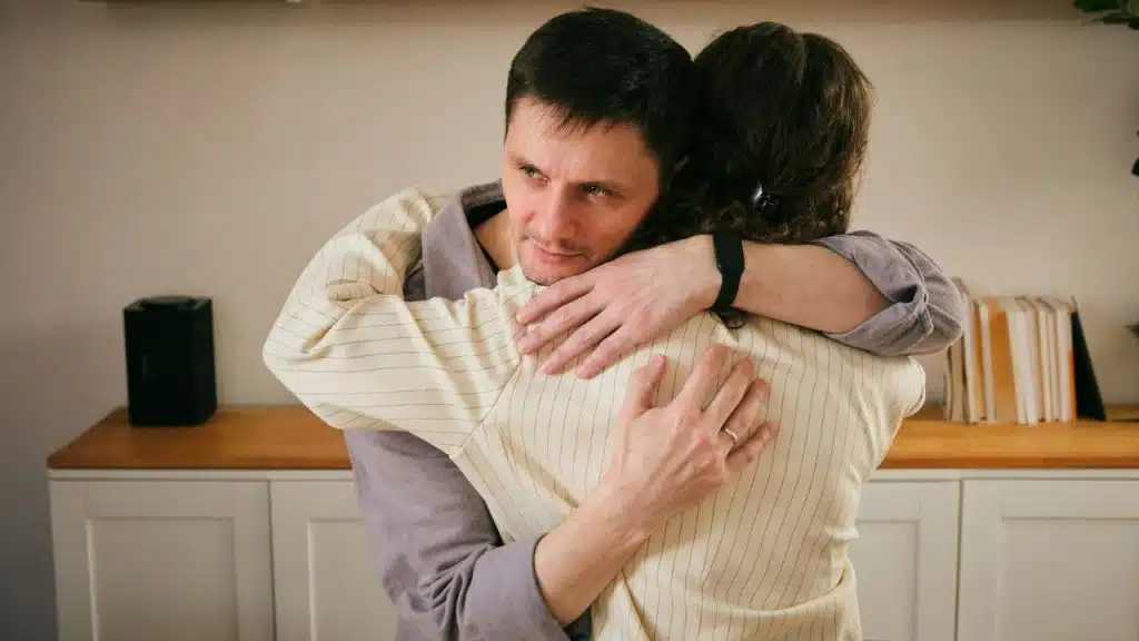 A man with a concerned expression hugs a person wearing a striped shirt indoors.