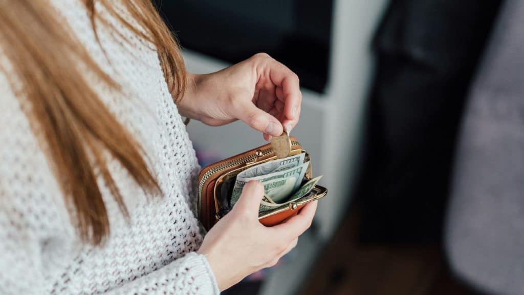 A person in a white sweater holds an open wallet containing cash and a coin.