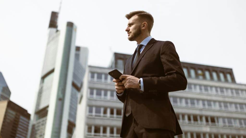A man in a dark suit holds a smartphone while standing in front of skyscrapers.